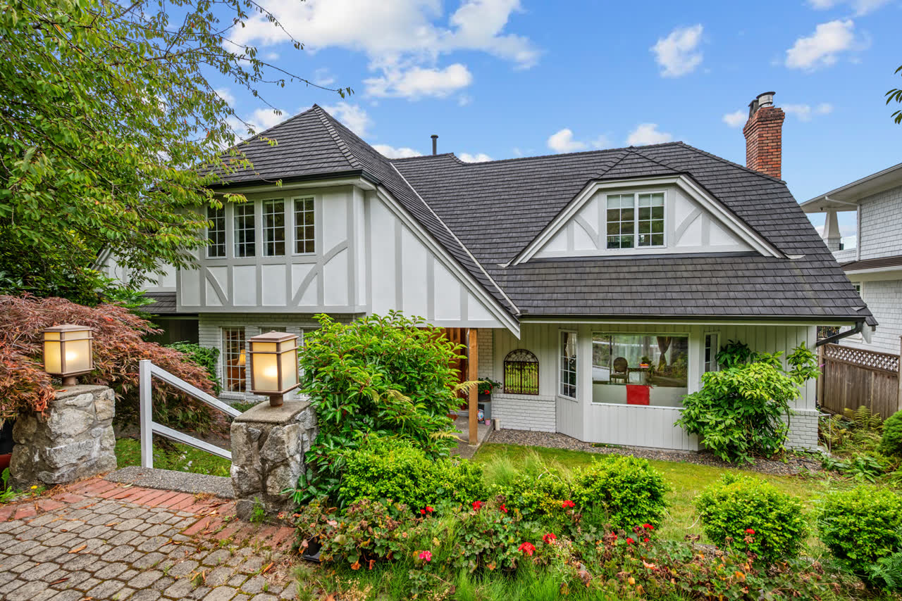 White Tudor-style home with a dark shingled roof, stone lantern posts and a brick paver path through flowering garden beds