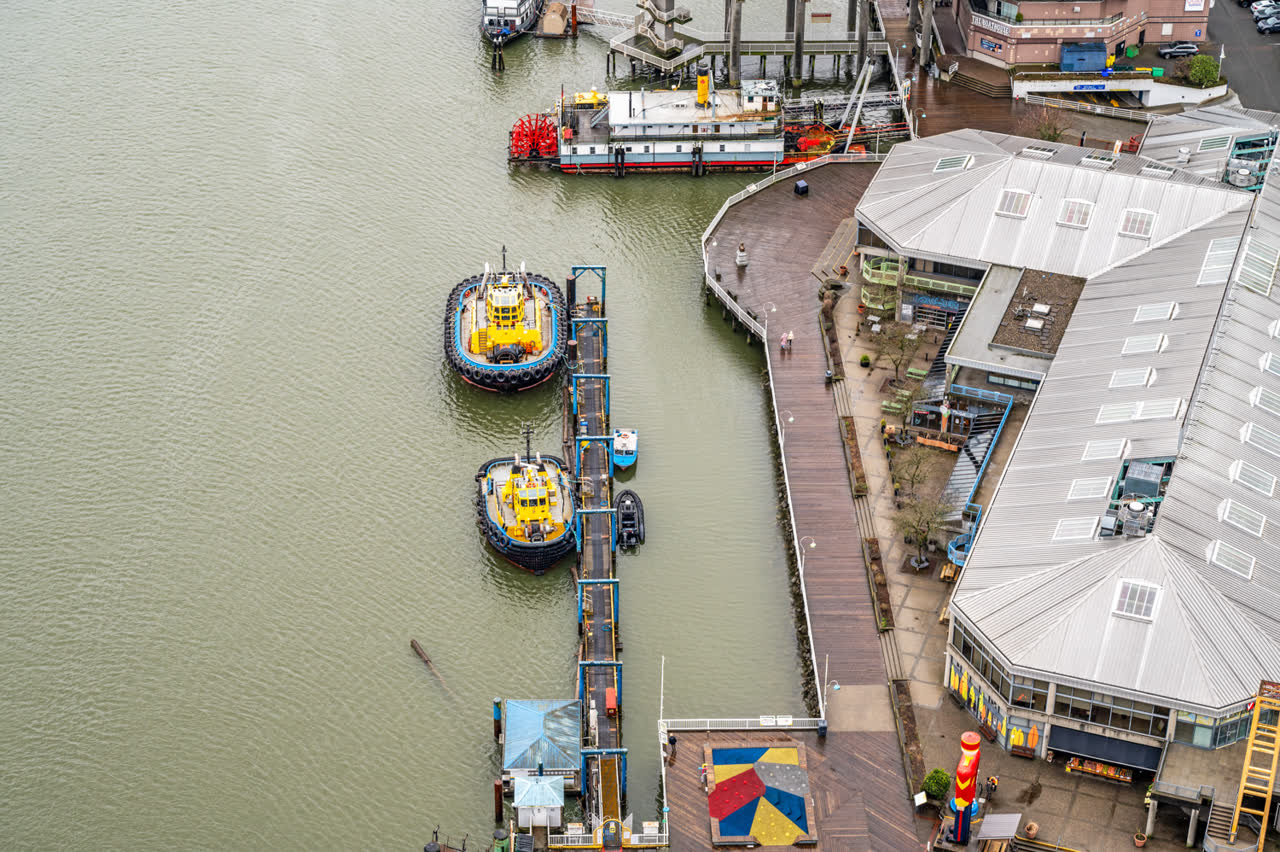 Aerial view of a waterfront marina, with moored tugboats alongside a covered market building and boardwalk